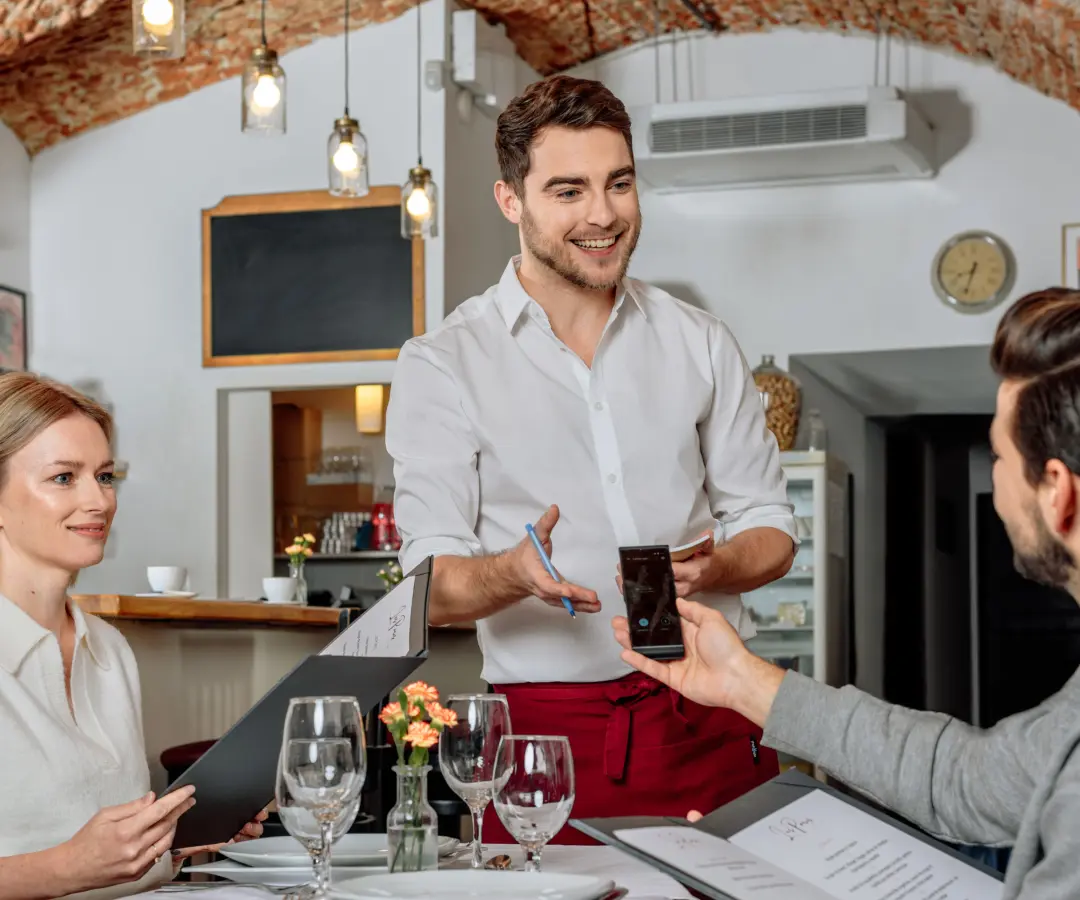 Un camarero sonriente está de pie junto a la mesa de un restaurante, mientras un cliente sentado sostiene un traductor en la mano. Una clienta se sienta cerca, con un menú en la mano. La mesa está puesta con vasos, platos y un pequeño jarrón de flores.