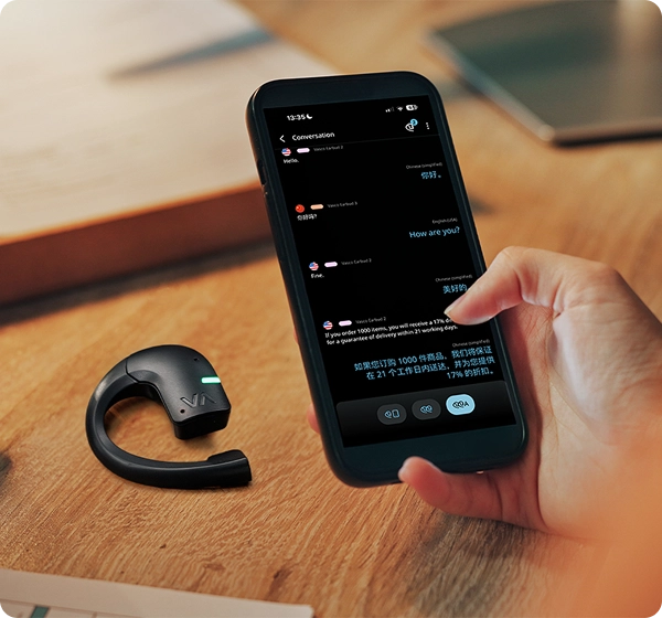 Primer plano de una mano que sostiene un smartphone en el que se muestra un chat con traducción en tiempo real entre inglés y chino. Junto al teléfono, sobre un escritorio de madera, descansa un auricular de traducción Vasco de color negro con un indicador LED verde que parpadea. 