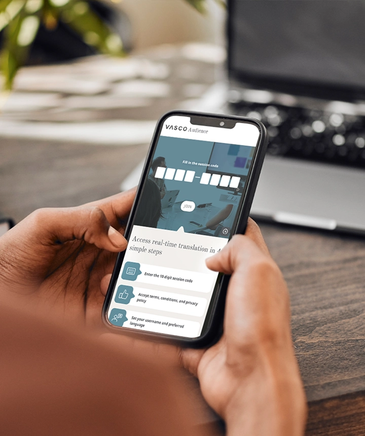 Una vista desde detrás de una persona que sostiene un smartphone en el que se muestra la interfaz web de Vasco Audience. La pantalla muestra un campo de entrada con el texto «Introduzca el código de sesión» y un botón blanco con la palabra «UNIRSE», situado encima de una lista de instrucciones que dice «Acceda a la traducción en tiempo real en 4 sencillos pasos». Al fondo se ve un escritorio de madera desenfocado y un ordenador portátil. 