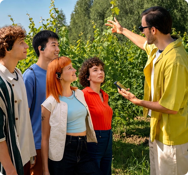Un grupo de cinco personas al aire libre en un viñedo bañado por el sol. A la derecha, un hombre con camisa amarilla y gafas de sol sostiene un dispositivo Translator Vasco Q1 y señala con un gesto las vides verdes. A la izquierda, cuatro personas escuchan con atención, cada una con un Translator Vasco E1 negro, lo que pone de manifiesto la función de conversación en grupo en tiempo real. 
