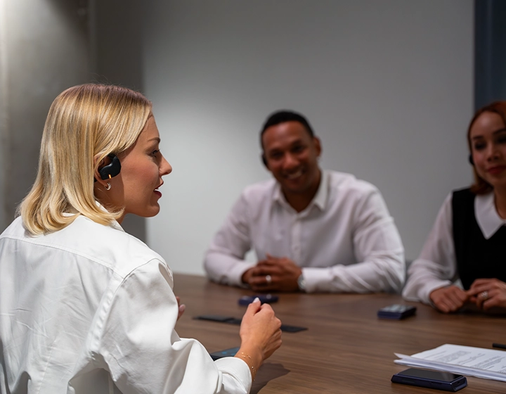 Perfil de una mujer rubia con una blusa blanca hablando durante una reunión de negocios en una mesa de conferencias de madera. Lleva puesto un auricular negro Vasco Translation E1. En el fondo, difuminado, hay otros dos profesionales sentados al otro lado de la mesa, sonriendo y escuchando con atención. 