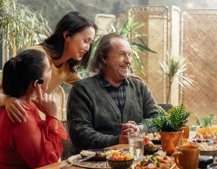 Una conmovedora reunión familiar intergeneracional alrededor de una mesa llena de comida. Un hombre mayor, sonriente y vestido con un cárdigan gris, y dos mujeres charlan alegremente en una habitación luminosa llena de plantas. Los tres llevan puestos los auriculares negros Vasco Translation E1. 
