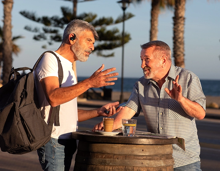 Dos hombres están de pie junto a una mesa al aire libre y conversan. Uno de ellos lleva una mochila y ambos utilizan los auriculares Vasco Translator E1. 