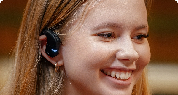 Primer plano del perfil de una joven sonriente que lleva un auricular Vasco Translation E1 negro. La imagen destaca el diseño higiénico y abierto del dispositivo, mostrando cómo se ajusta cómodamente en la parte exterior de la oreja sin introducirse en el conducto auditivo. 