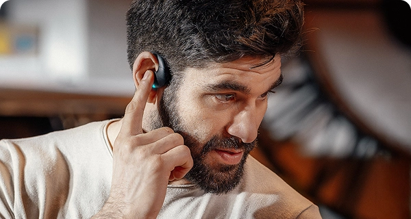 Primer plano de un hombre barbudo que toca con el dedo índice un auricular negro de traducción Vasco que lleva en la oreja, mostrando así sus intuitivos controles táctiles. En el dispositivo se ve encendida una pequeña luz LED de color azul brillante. 