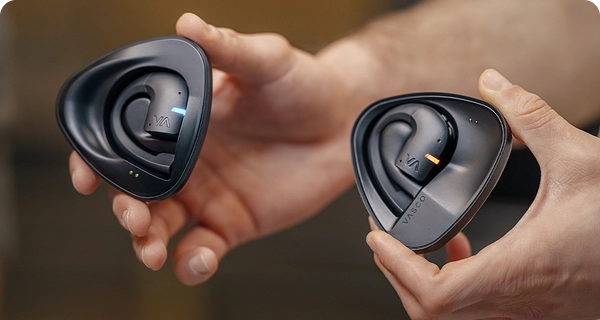 Primer plano de unas manos que sostienen dos estuches de carga negros independientes con los auriculares de traducción Vasco. La imagen muestra cómo distinguir ambos estuches: uno tiene un LED azul y el otro, uno naranja, que indican los distintos altavoces. 