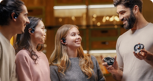 Un grupo de cuatro personas de diferentes procedencias muestra el diseño universal de los auriculares Vasco Translation E1 en una habitación con una iluminación cálida. Tres de ellas llevan puestos los auriculares negros, demostrando que se adaptan cómodamente a diferentes formas de oreja, mientras que un hombre barbudo y sonriente, situado a la derecha, sostiene dos estuches de carga. 
