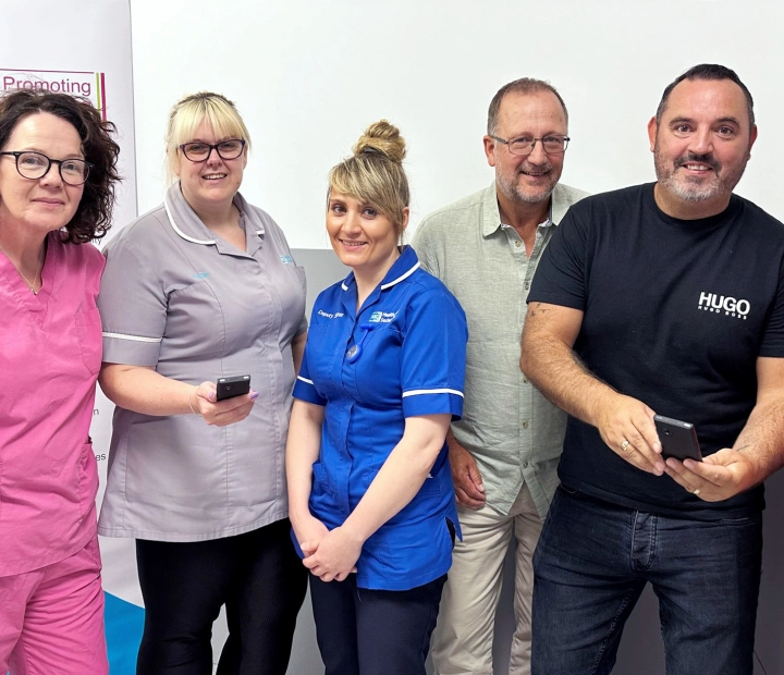 Cinco personas sonrientes están de pie juntos, 3 vestidos con ropa sanitaria. Dos de ellos sostienen un dispositivo traductor.