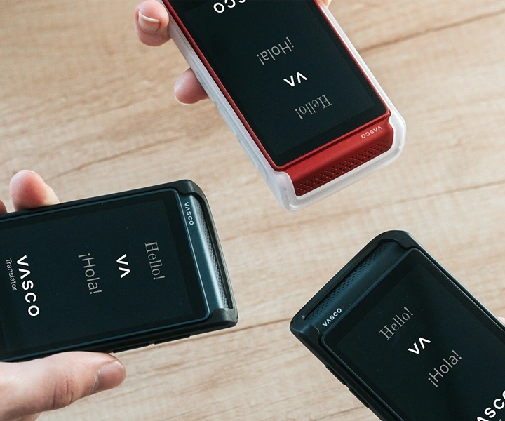 Tres personas que sostienen el Traductor Q1 los juntan; las pantallas muestran la pantalla de inicio con el logotipo de Vasco y las palabras «Hello!» y «¡Hola!». Los dispositivos se presentan en fundas de silicona en tres colores: Solid Black, Dark Caramel, and Milky White.