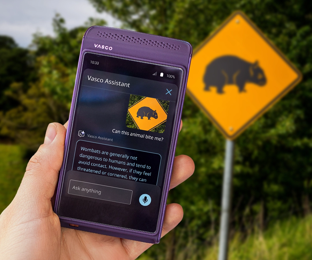 Una mano sosteniendo un traductor VA de color morado. En la pantalla del dispositivo se ve la aplicación Vasco Assistant, que muestra una foto de una señal de advertencia con la imagen de un wombat y una descripción sobre estos animales. Al fondo, detrás de la mano que sostiene el dispositivo, se ve una carretera y la misma señal de tráfico amarilla con un wombat que aparece en la pantalla del traductor.
